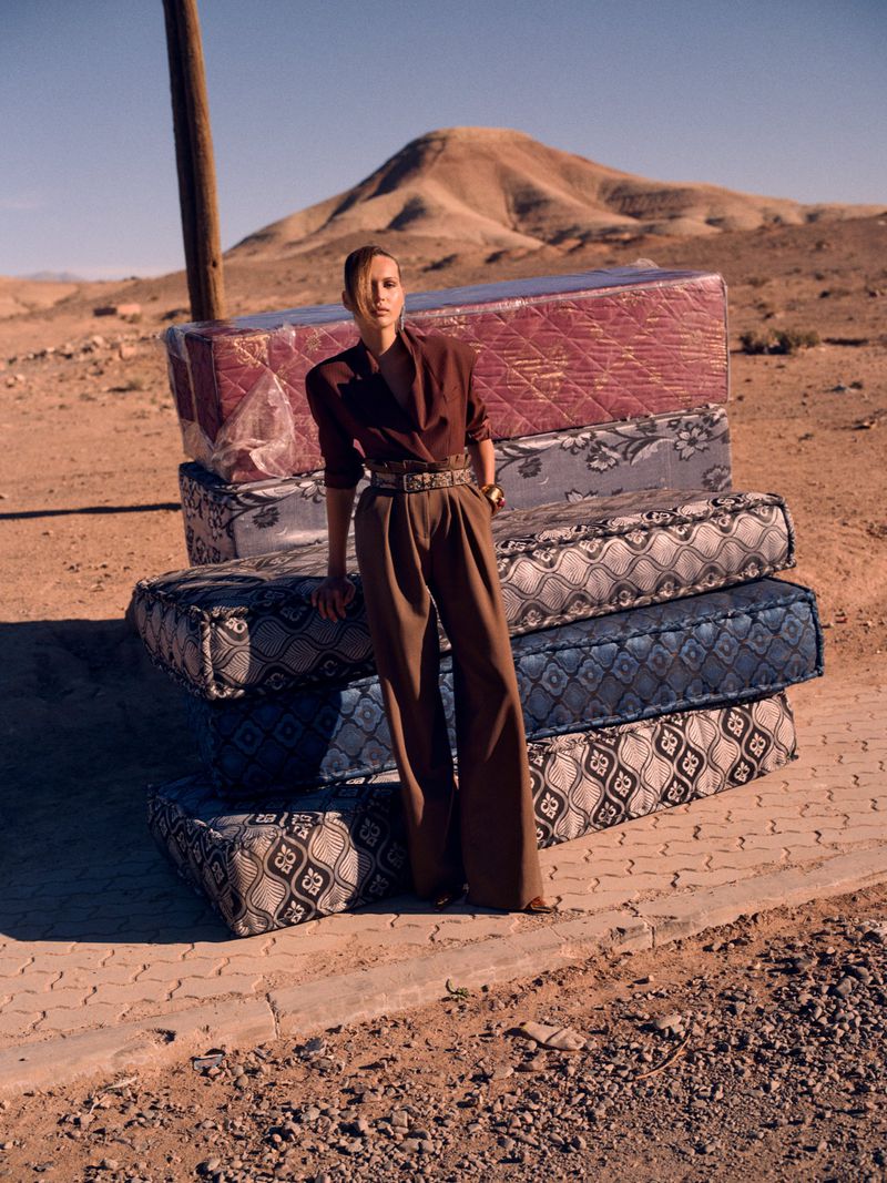 Woman posing on a stack of mattresses in the desert, wearing a brown blouse and pants. A mountain is visible in the background.