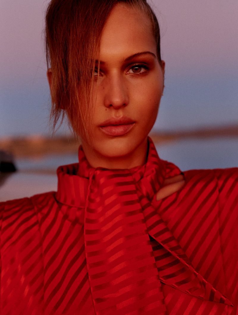 Close-up portrait of a woman with red hair and red striped dress against a soft sky background. Fashion editorial style, beauty shot.