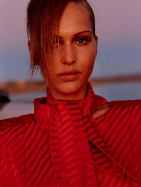 Close-up portrait of a woman with red hair and red striped dress against a soft sky background. Fashion editorial style, beauty shot.