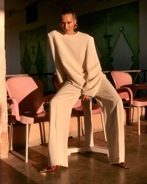 Fashionable woman in cream wide-leg pants and a matching long-sleeve top posing in an outdoor setting with pink chairs.