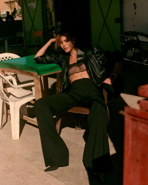 Fashion shot of a brunette woman in an outdoor setting, posing on chair with an open black leather jacket and wide-leg black pants.