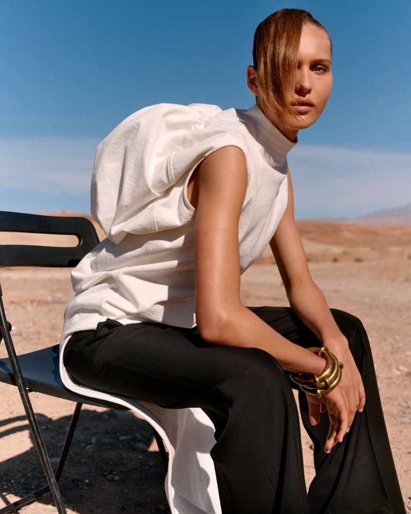 A woman in a white top and black pants sits on a chair in a desert landscape under a clear blue sky, showcasing fashion and style.