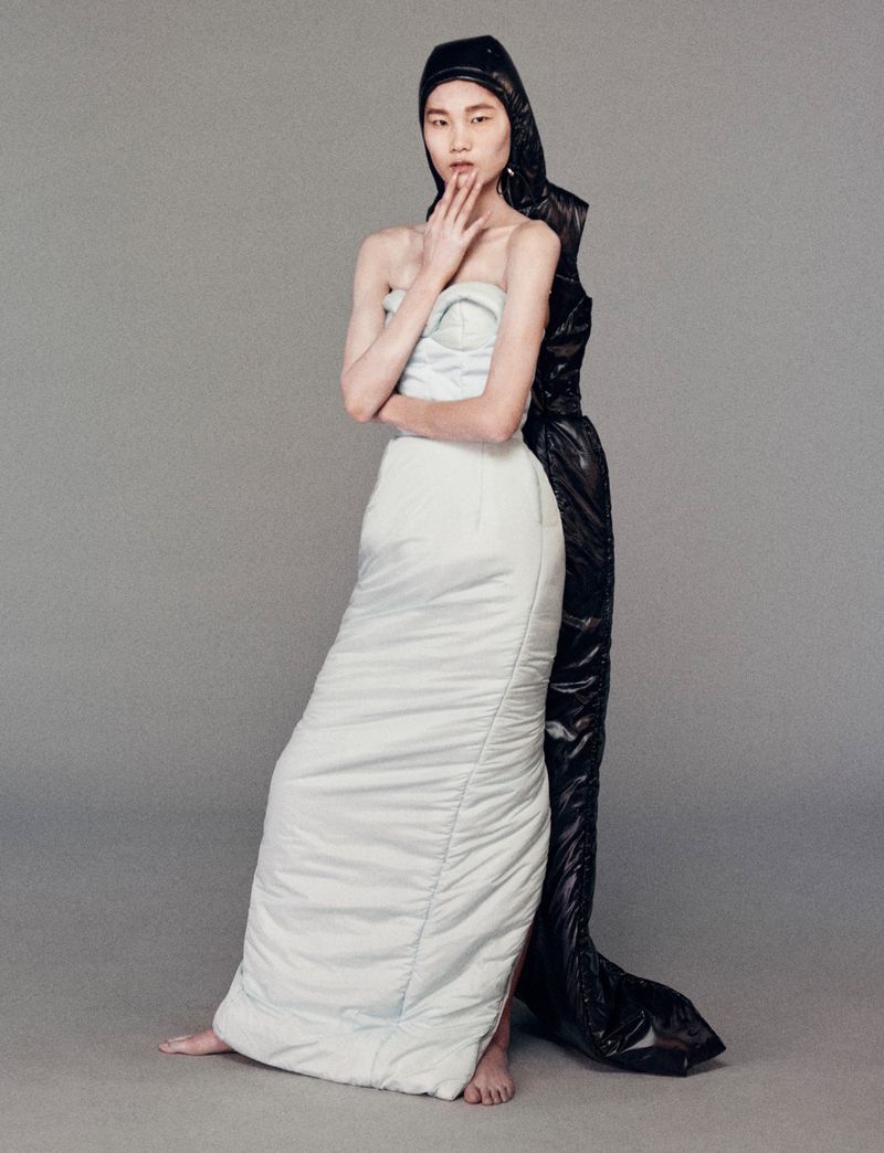 Two fashion models in a photo studio pose against a grey backdrop. One wears a white padded dress, and the other a black hooded jumpsuit.