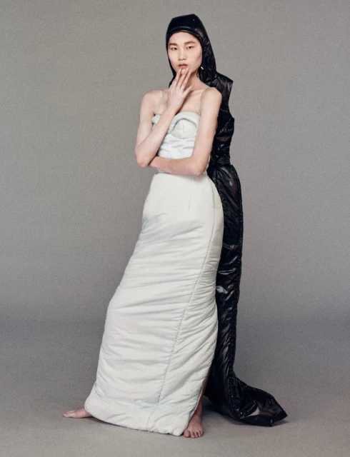Two fashion models in a photo studio pose against a grey backdrop. One wears a white padded dress, and the other a black hooded jumpsuit.
