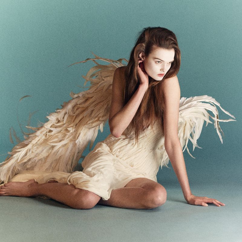 Young woman in a white dress and feather wings poses against a blue background in a studio portrait showcasing fashion and fine art.