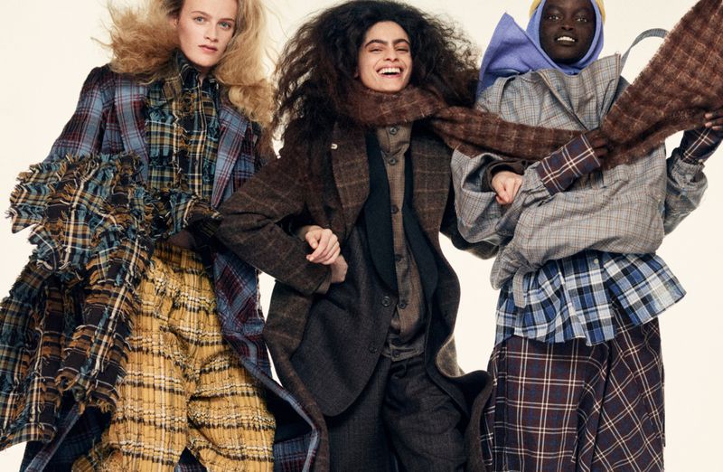Three diverse fashion models wearing colorful plaid and tweed coats, skirts, and scarves pose together in a studio setting.