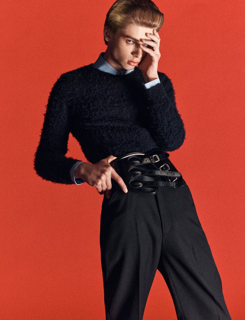 A young adult male poses in front of an orange backdrop, wearing a navy sweater, black pants, and a multi-belted leather corset.