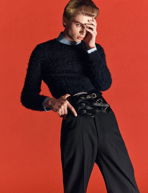 A young adult male poses in front of an orange backdrop, wearing a navy sweater, black pants, and a multi-belted leather corset.