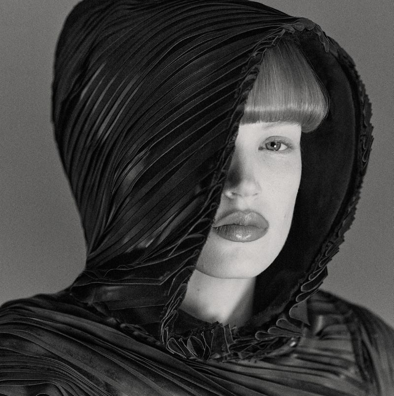 Monochrome portrait of a female fashion model wearing an elaborate, textured black hood that partially covers her face in a studio setting.
