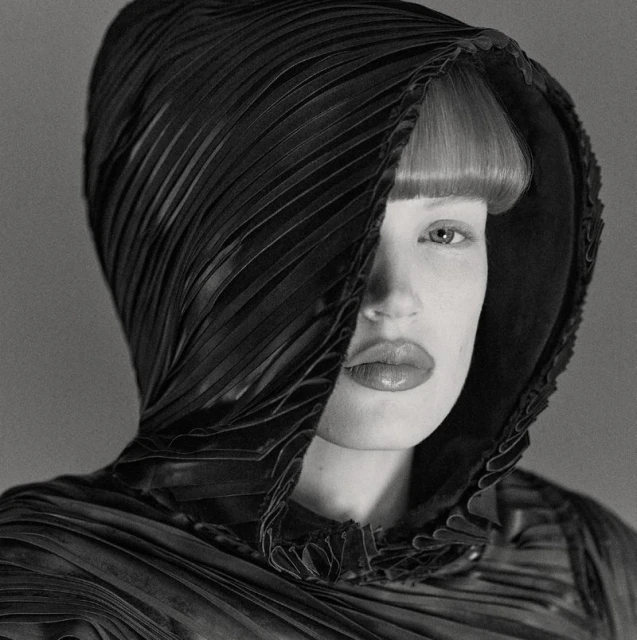 Monochrome portrait of a female fashion model wearing an elaborate, textured black hood that partially covers her face in a studio setting.