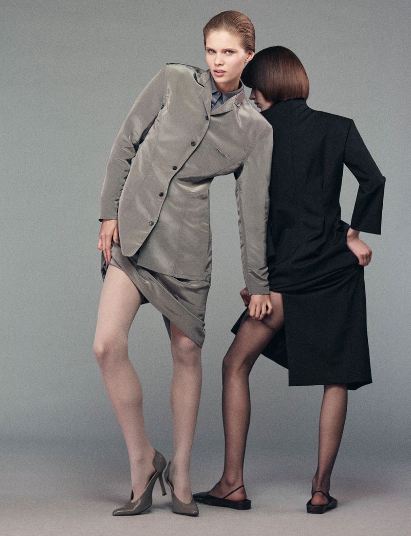 Two women pose in a fashion shot in a studio with neutral background. One woman wears a grey suit and heels, the other a black dress and flats.