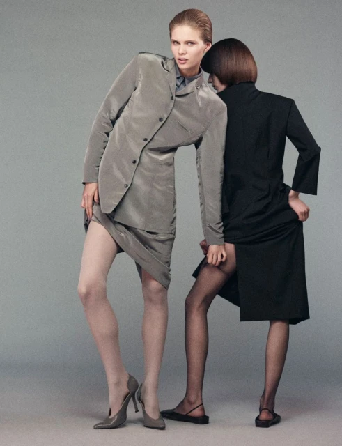 Two women pose in a fashion shot in a studio with neutral background. One woman wears a grey suit and heels, the other a black dress and flats.