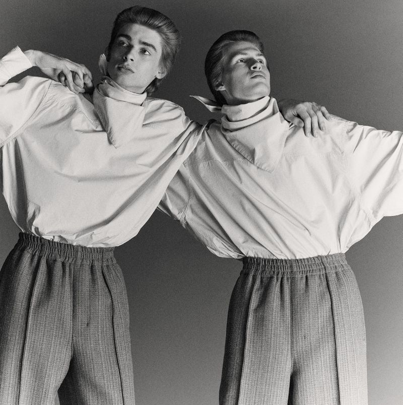 Two adult men in matching outfits pose in a studio. They wear button-down shirts with neckerchiefs and high-waisted trousers with elastic waistbands.