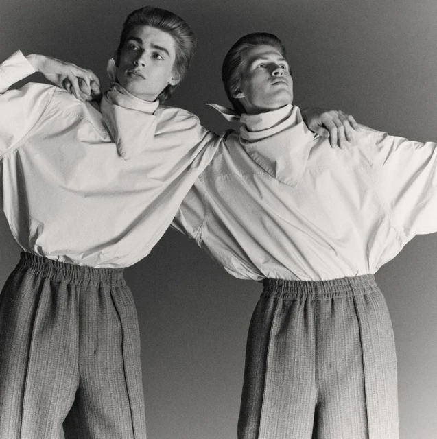 Two adult men in matching outfits pose in a studio. They wear button-down shirts with neckerchiefs and high-waisted trousers with elastic waistbands.