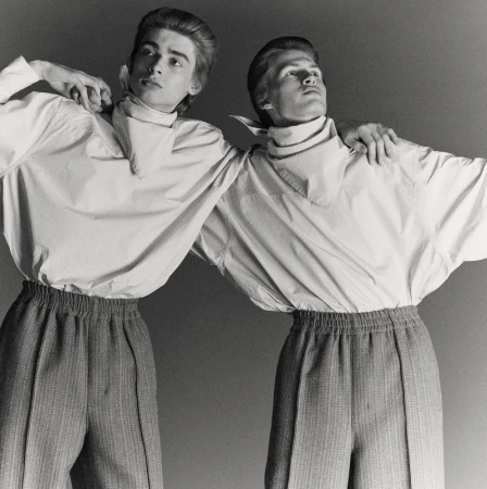 Two adult men in matching outfits pose in a studio. They wear button-down shirts with neckerchiefs and high-waisted trousers with elastic waistbands.