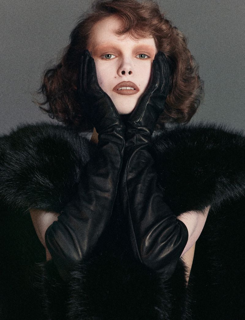 Close-up portrait of a woman with pale skin, wearing a black fur coat and gloves, posing with her hands near her face.