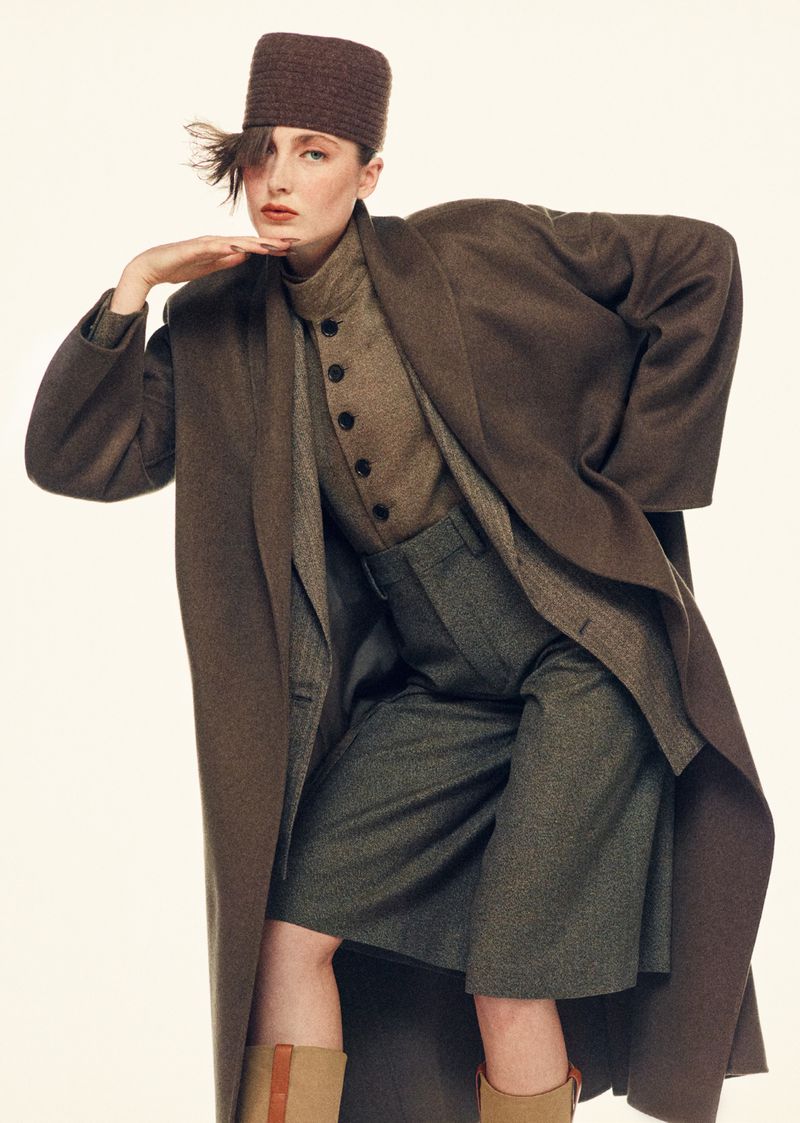 A stylish female fashion model in a brown hat and long brown coat poses in a studio with a neutral background for a fashion editorial.