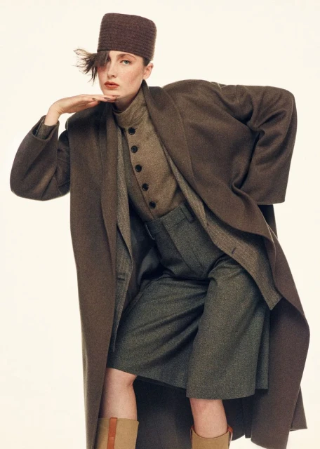 A stylish female fashion model in a brown hat and long brown coat poses in a studio with a neutral background for a fashion editorial.