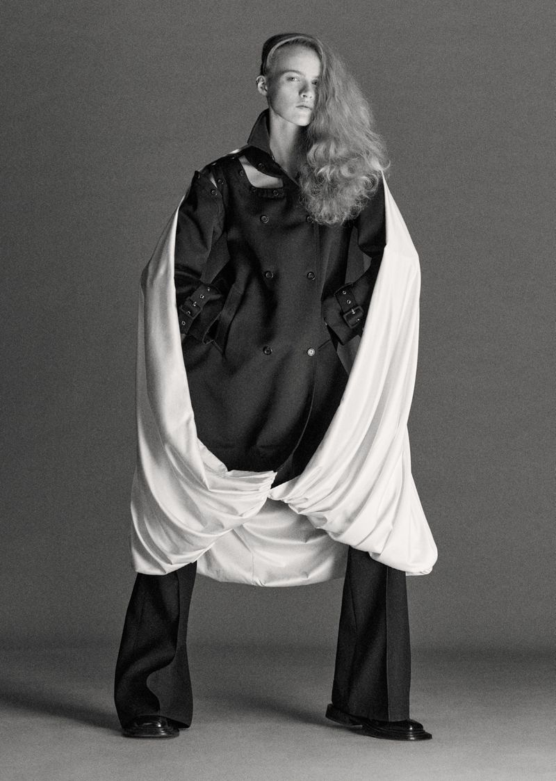 Fashion portrait of a young fair-skinned woman with long blond hair in a black coat with cream fabric detail and black pants against a gray background.