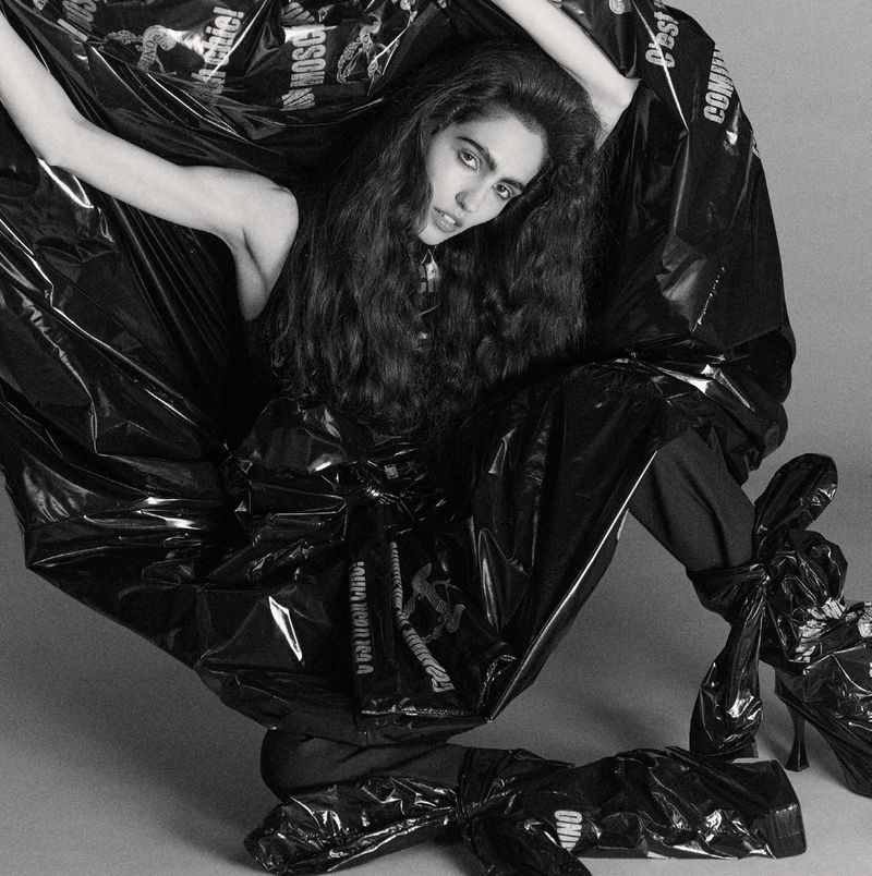 Woman in a black plastic outfit with designer logos poses in a studio. Fashion photography with a minimalist background and creative costuming.