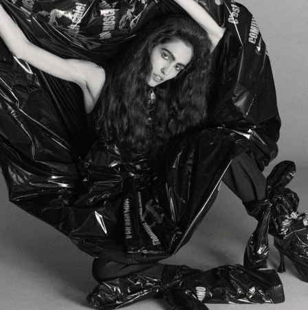 Woman in a black plastic outfit with designer logos poses in a studio. Fashion photography with a minimalist background and creative costuming.