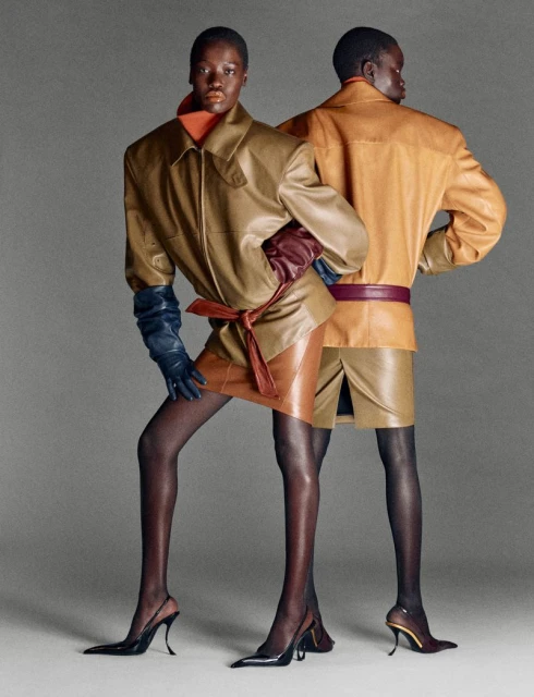 Two dark-skinned fashion models pose in tan leather jackets and skirts on a neutral backdrop, with high-heeled shoes and colored leather gloves.