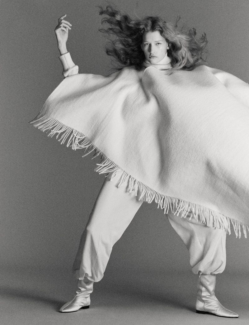 Monochrome portrait of a model in motion wearing a poncho, baggy pants, and silver boots against a grey backdrop.