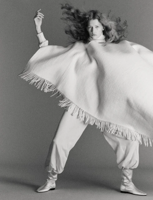 Monochrome portrait of a model in motion wearing a poncho, baggy pants, and silver boots against a grey backdrop.