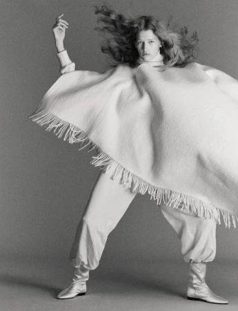 Monochrome portrait of a model in motion wearing a poncho, baggy pants, and silver boots against a grey backdrop.