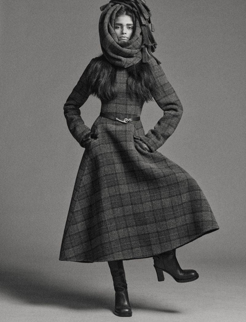 Monochrome studio shot of a fashion model wearing a plaid coat and boots with a scarf wrapped around the head.