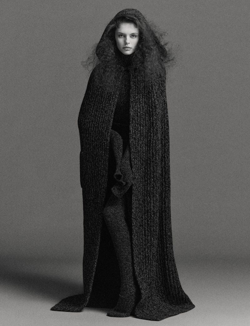 A solitary adult female is captured in a studio shot, draped in a hooded, dark gray knitted cape dress, paired with matching stocking boots.