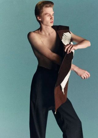 Fashion shot of a young, fair-skinned male model in black pants, wearing a brown and white vest-like garment, posing against a blue background.