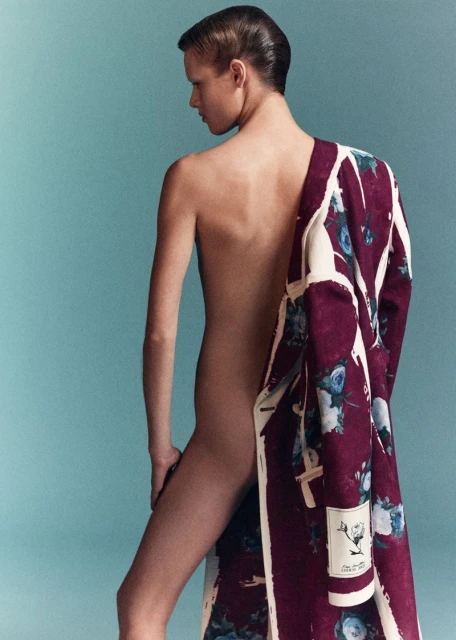 Torso of a slender person with short slicked-back hair, partially draped in a patterned maroon and cream coat, against a light blue backdrop.