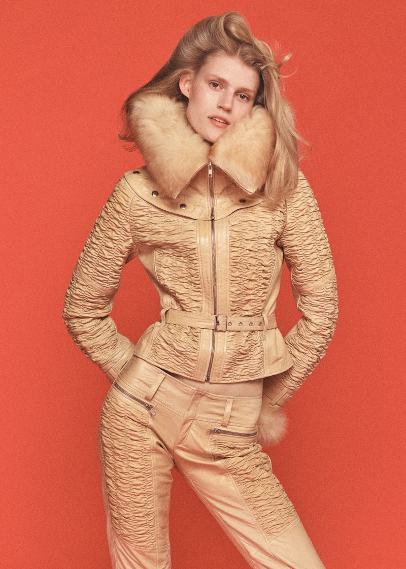 A woman poses in a tan, form-fitting jacket and pants with fur trim against a red backdrop. The jacket is belted, with matching pants.