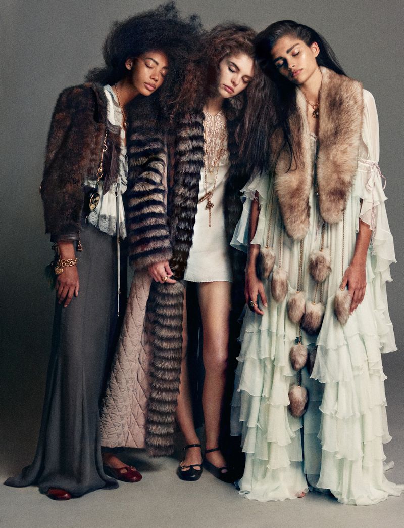 Three fashion models in tiered dresses and fur coats pose in a studio with neutral grey backdrop. Editorial fashion photography.