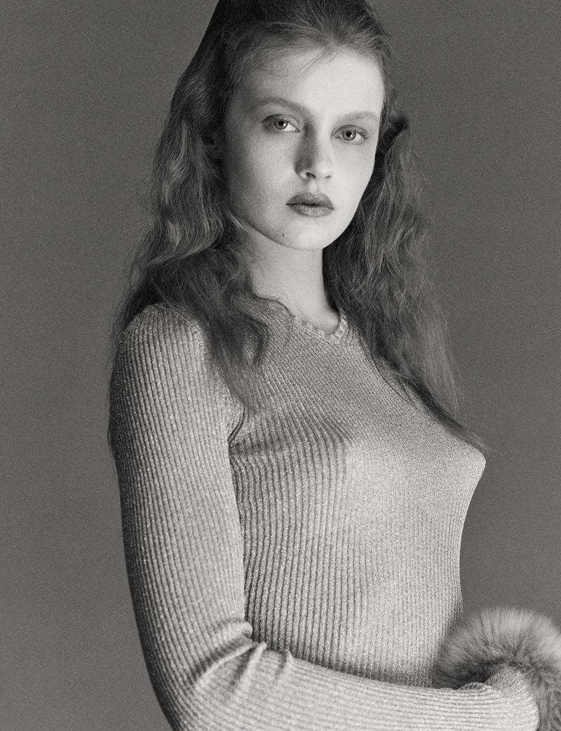 Monochrome portrait of a young adult woman with wavy hair, wearing a long-sleeved top with fur cuffs against a grey backdrop in a photo studio.