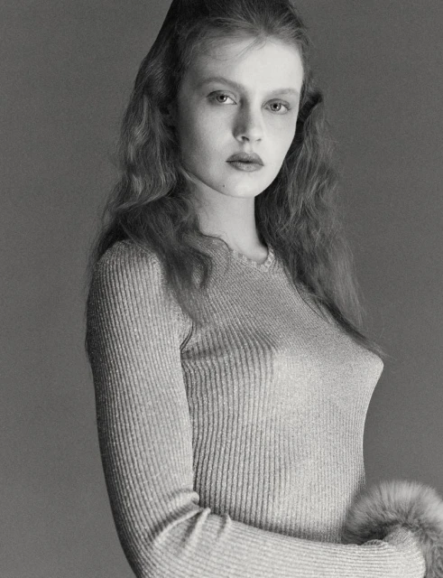Monochrome portrait of a young adult woman with wavy hair, wearing a long-sleeved top with fur cuffs against a grey backdrop in a photo studio.
