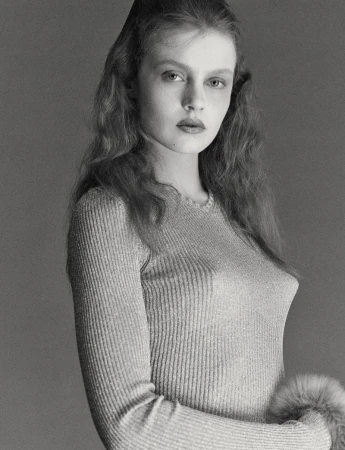 Monochrome portrait of a young adult woman with wavy hair, wearing a long-sleeved top with fur cuffs against a grey backdrop in a photo studio.