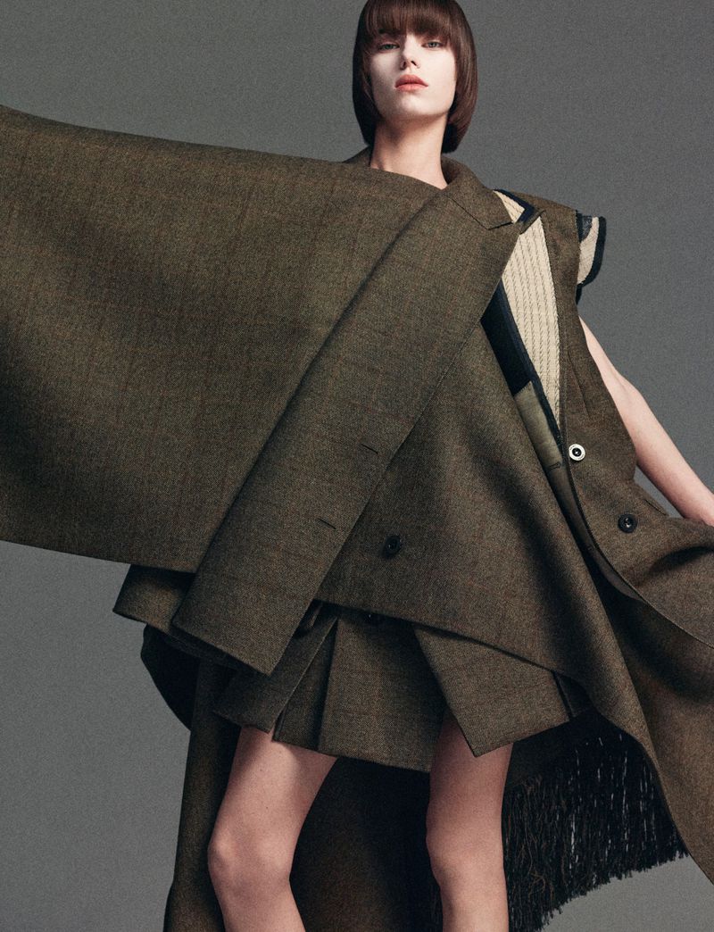 Editorial fashion shot of a woman wearing an oversized, unstructured brown jacket and pleated skirt in front of a gray backdrop.
