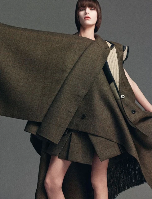 Editorial fashion shot of a woman wearing an oversized, unstructured brown jacket and pleated skirt in front of a gray backdrop.