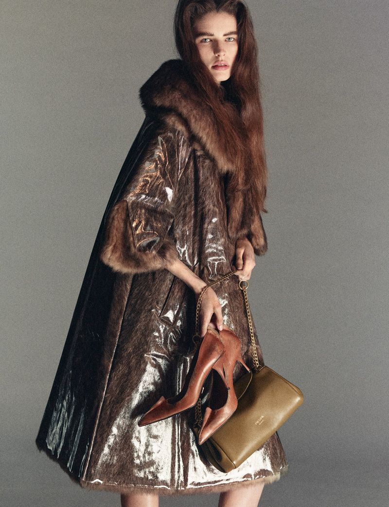 Fashionable woman in brown fur-trimmed coat holds brown heels and purse against a neutral background in a studio shot.