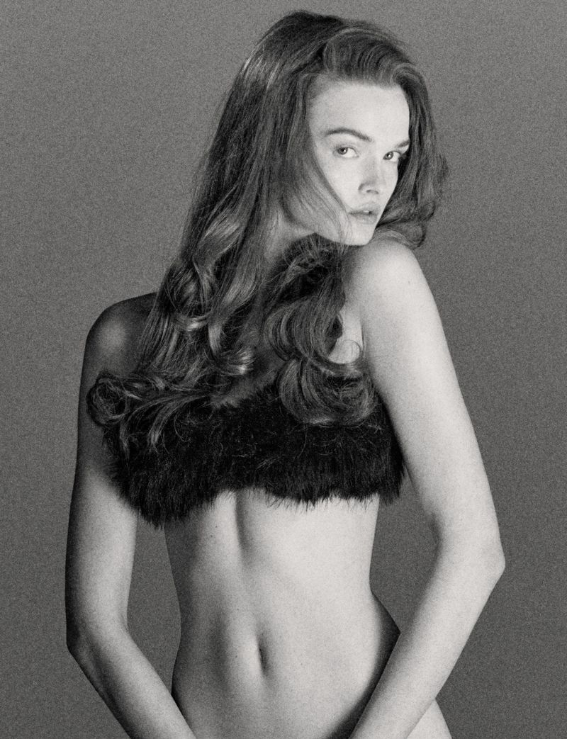 Monochrome portrait of a woman with long, wavy hair, posing with a black, fur-like garment. Studio shot with a grey backdrop.