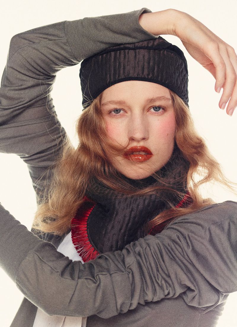 A young adult female fashion model wearing a gray turtleneck and matching cap poses in a studio with a bright white background.