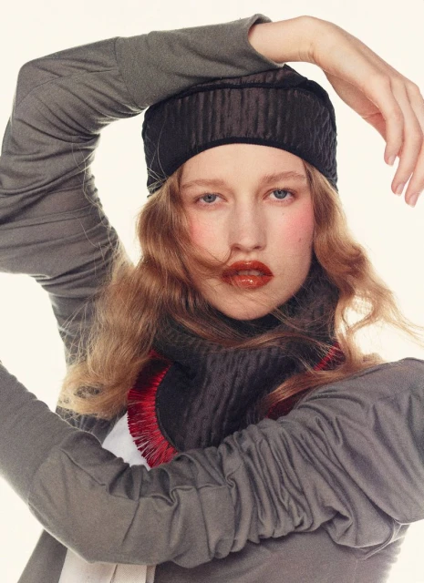 A young adult female fashion model wearing a gray turtleneck and matching cap poses in a studio with a bright white background.