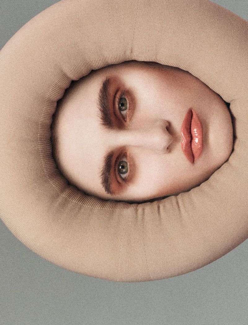 A surreal, close-up portrait of a person's face framed by a large, rounded, cushioned collar in a studio setting with neutral lighting.