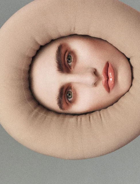 A surreal, close-up portrait of a person's face framed by a large, rounded, cushioned collar in a studio setting with neutral lighting.