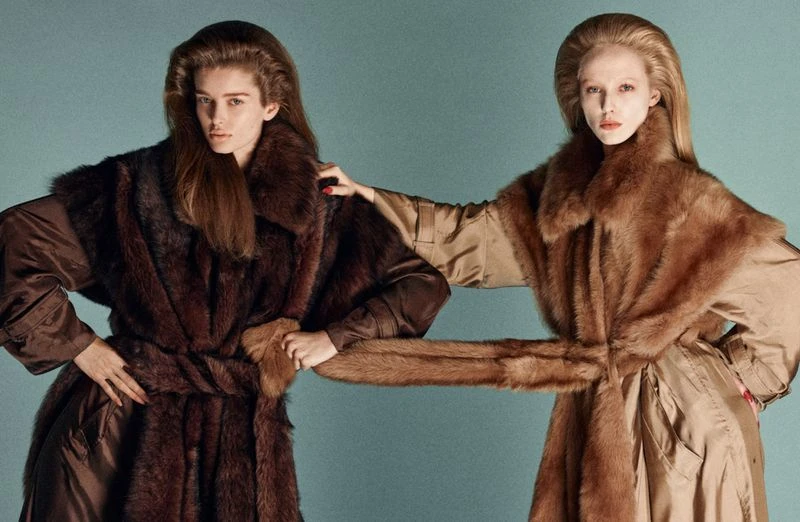Two fashion models pose in coordinated brown fur coats against a neutral background, showcasing luxury and style in a studio setting.