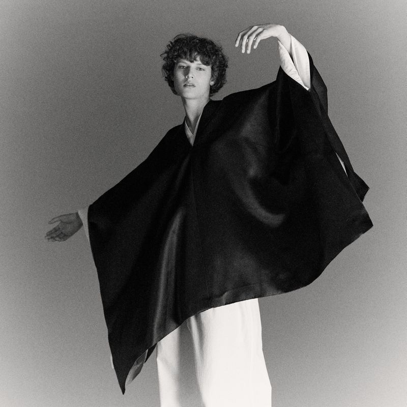 Monochrome studio shot of a slender person with curly hair in an elegant draped black garment, striking a dynamic pose with arms outstretched.