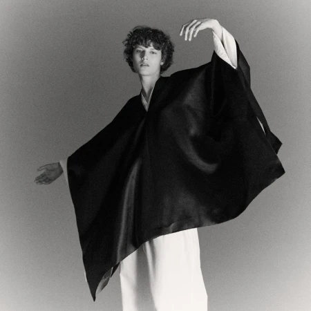 Monochrome studio shot of a slender person with curly hair in an elegant draped black garment, striking a dynamic pose with arms outstretched.