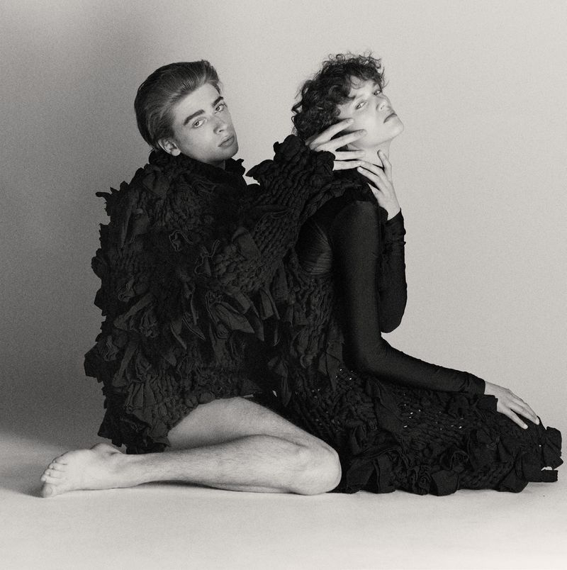Two people pose in black clothing against a light background in a studio shot. One person is sitting with their hand touching the other person.